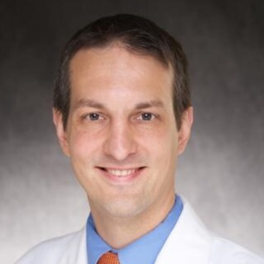 A photo of Dr. Aaron Boes wearing a white coat, blue shirt, and tie in front of a gray background