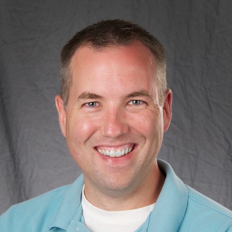 A photo of Joel Bruss smiling, wearing a blue shirt in front of a gray background